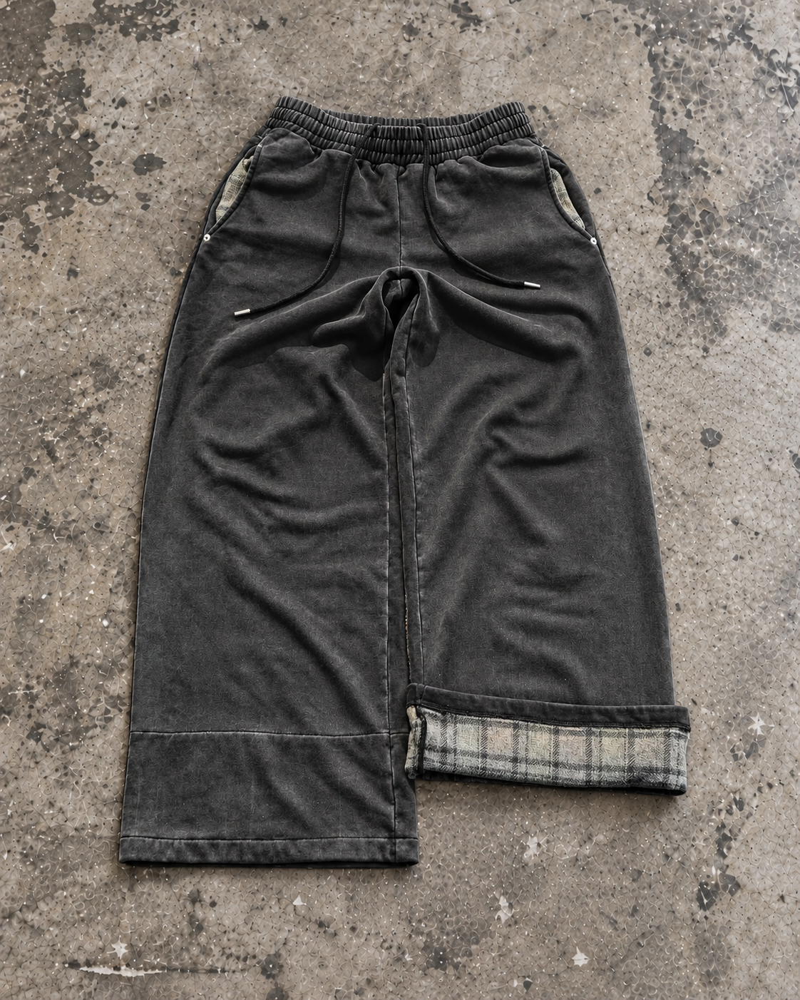 Flannel-lined wide pants in washed black with plaid cuff, flat lay on concrete